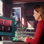 A woman in a red shirt sits at a desk using a computer, editing or reviewing images of a person in different poses. The workspace has office supplies and a modern, exposed brick wall background.