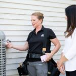 A technician in a black polo shirt and tool belt explains something about an outdoor AC unit to a woman in a white jacket, both standing next to a house with white siding.