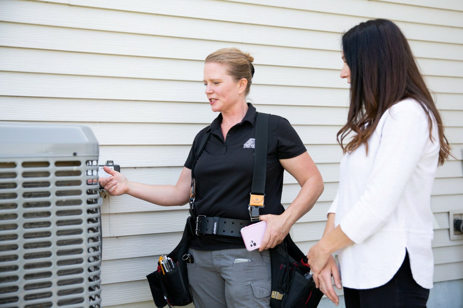 A technician in a black polo shirt and tool belt explains something about an outdoor AC unit to a woman in a white jacket, both standing next to a house with white siding.