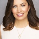 A woman with wavy brown hair, green eyes, and a bright smile is wearing a sleeveless white top, pearl stud earrings, and a gold necklace with a heart-shaped pendant, against a neutral gray background.