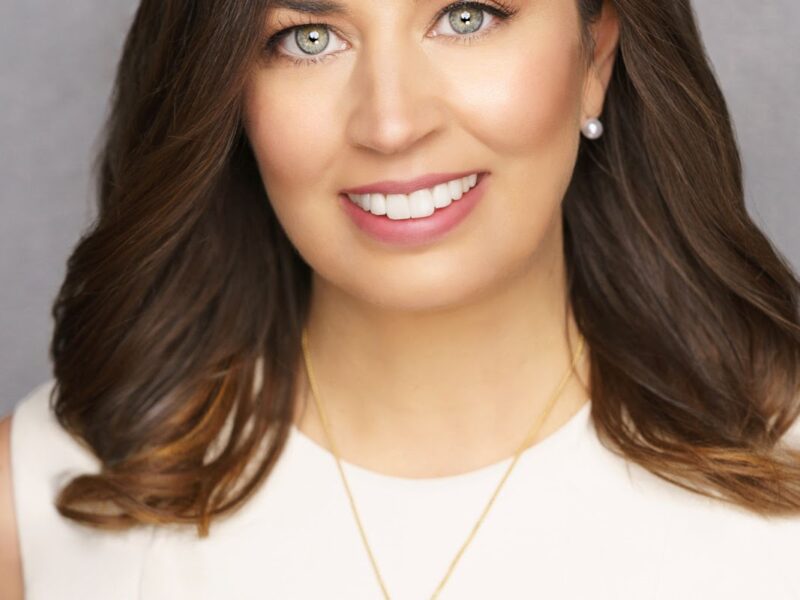A woman with wavy brown hair, green eyes, and a bright smile is wearing a sleeveless white top, pearl stud earrings, and a gold necklace with a heart-shaped pendant, against a neutral gray background.