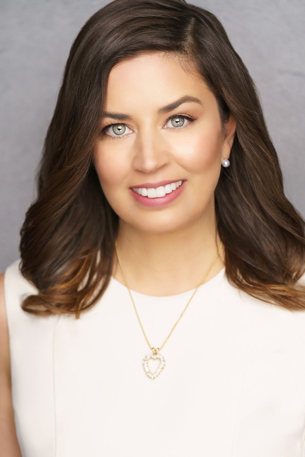 A woman with wavy brown hair, green eyes, and a bright smile is wearing a sleeveless white top, pearl stud earrings, and a gold necklace with a heart-shaped pendant, against a neutral gray background.