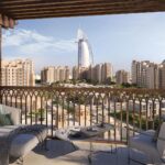 A balcony of Madinat Jumeirah with modern outdoor furniture overlooks a cityscape of beige buildings, featuring the iconic Burj Al Arab hotel in the distance under a clear blue sky.