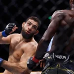Two mixed martial artists fight inside a cage. The shirtless fighter on the left is recoiling from a punch thrown by the shirtless fighter on the right, who is wearing black Venum shorts.