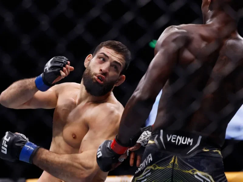 Two mixed martial artists fight inside a cage. The shirtless fighter on the left is recoiling from a punch thrown by the shirtless fighter on the right, who is wearing black Venum shorts.