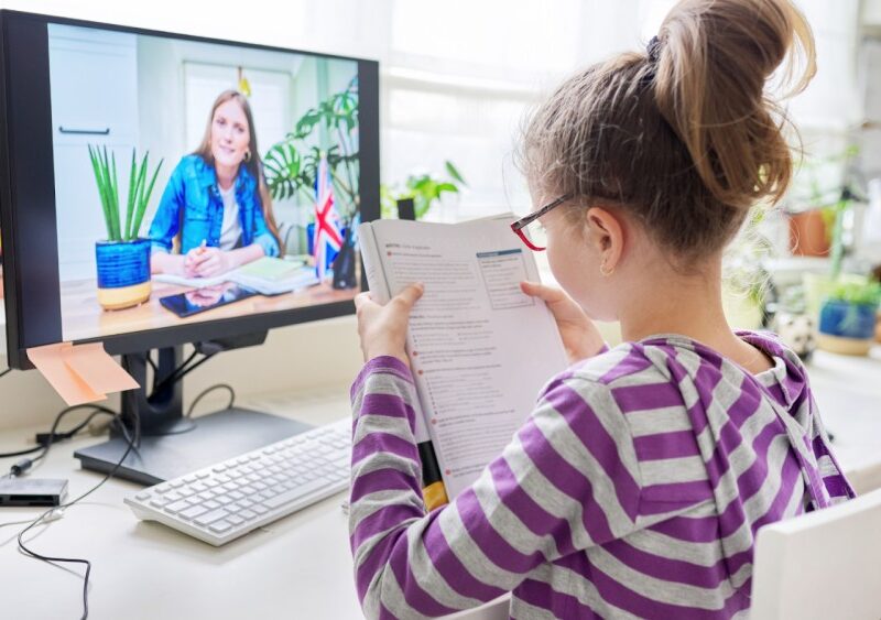 A young girl in a striped hoodie is sitting at a desk, holding an open book and attending an online class on a computer, with her teacher visible on the screen.