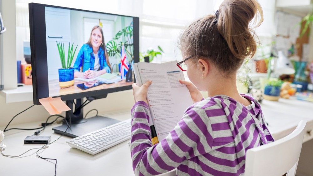 A young girl in a striped hoodie is sitting at a desk, holding an open book and attending an online class on a computer, with her teacher visible on the screen.