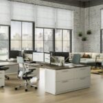 Modern, spacious office with large windows, white brick walls, light wood floors, and multiple desks with computers. There are ergonomic chairs, lounge seating, and plants adding a touch of greenery.