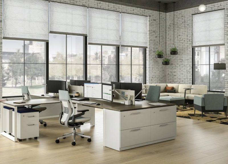 Modern, spacious office with large windows, white brick walls, light wood floors, and multiple desks with computers. There are ergonomic chairs, lounge seating, and plants adding a touch of greenery.