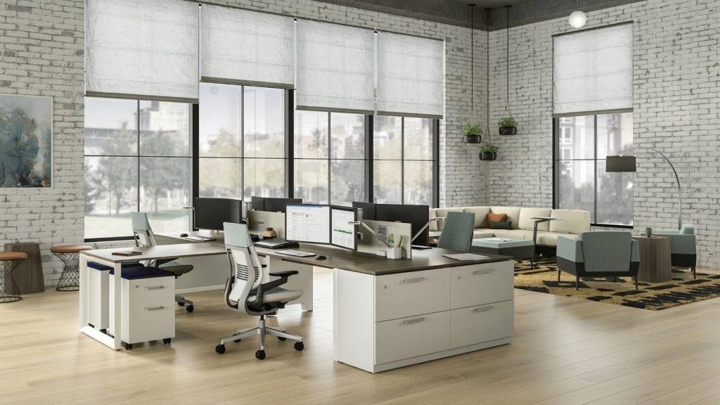 Modern, spacious office with large windows, white brick walls, light wood floors, and multiple desks with computers. There are ergonomic chairs, lounge seating, and plants adding a touch of greenery.