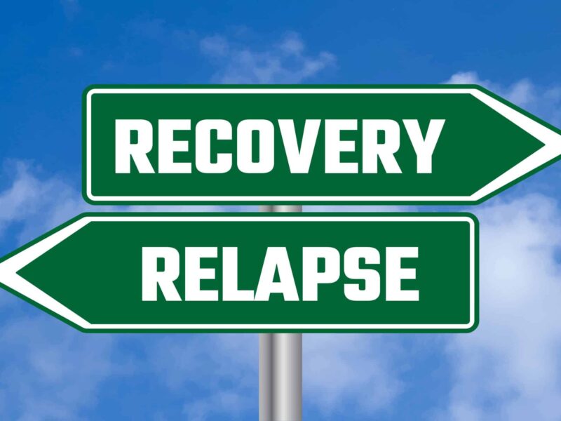 Two green directional signs against a blue sky with clouds; one points right and reads RECOVERY, the other points left and reads RELAPSE.