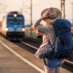 A person with a large blue backpack and a hat stands on a train platform, facing an approaching blue train, with one hand holding their hat and the other by their side. The scene appears to be during sunset.