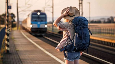 A person with a large blue backpack and a hat stands on a train platform, facing an approaching blue train, with one hand holding their hat and the other by their side. The scene appears to be during sunset.