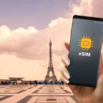 A hand holds a smartphone displaying an eSIM icon, with the Eiffel Tower and a large open plaza in Paris visible in the background under a partly cloudy sky.
