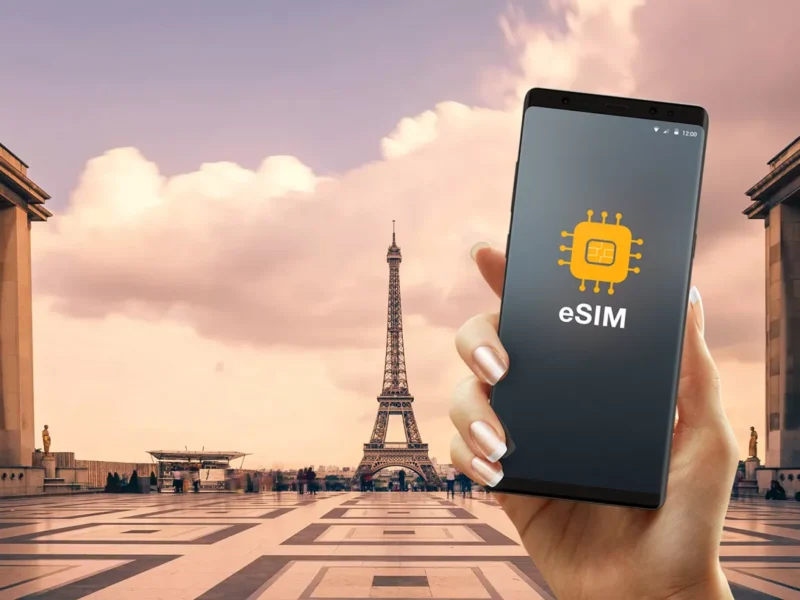A hand holds a smartphone displaying an eSIM icon, with the Eiffel Tower and a large open plaza in Paris visible in the background under a partly cloudy sky.